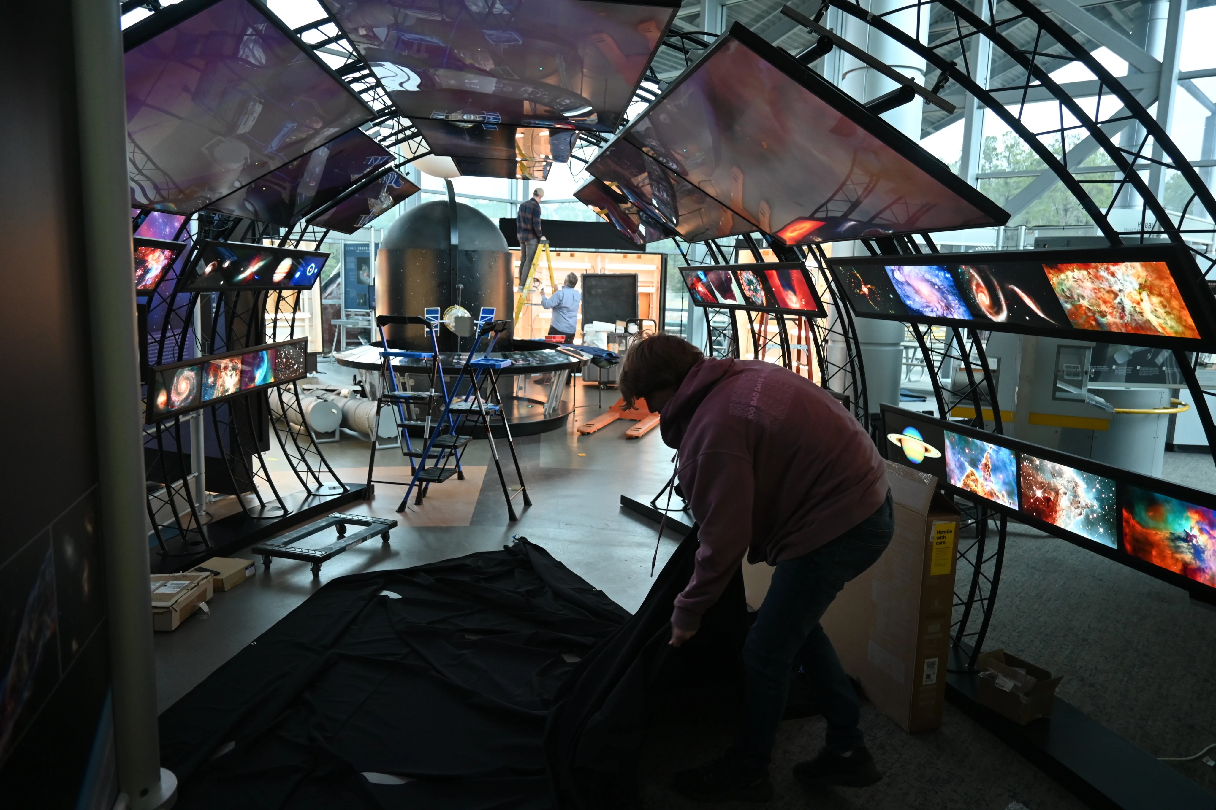 Several workers are seen assembling the Hubble traveling exhibit as various stations are in different states of completion.