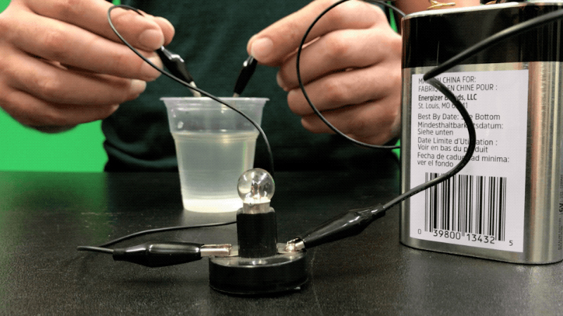 A person is shown using electricity, two cords, and a cup of water to turn on a mini light bulb.