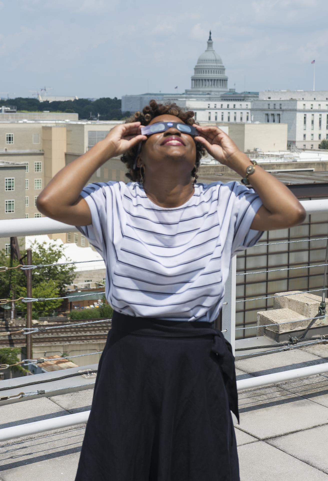 A woman stands on a rooftop, wearing eclipse classes, staring up at the sky.