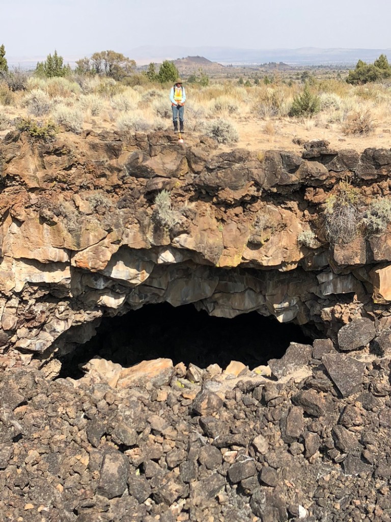 A researcher stands on a cliff edge. In the cliff face below, a dark cave opening is visible.