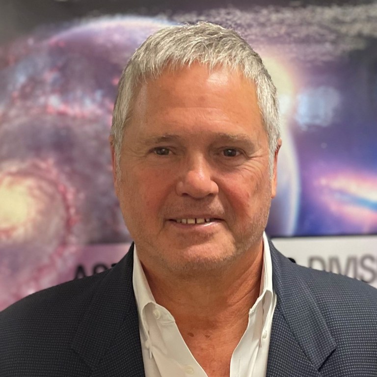 Headshot of Mr. Joe Smith wearing a white collared shirt and dark jacket, standing in front of a purple space poster.