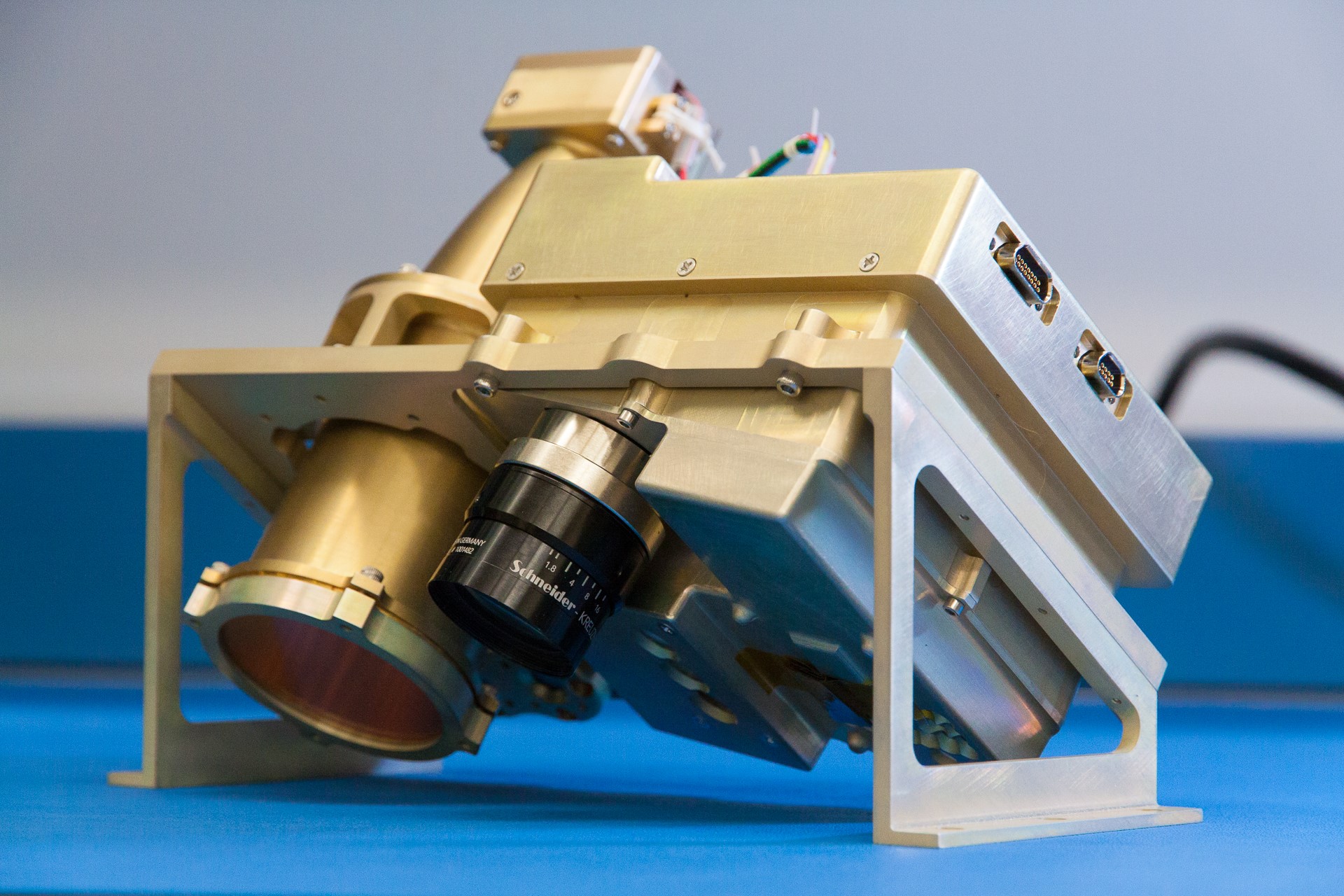 A gold-colored metal instrument with multiple cameras mounted pointing diagonally downwards. Two parallel ports are mounted on the side.