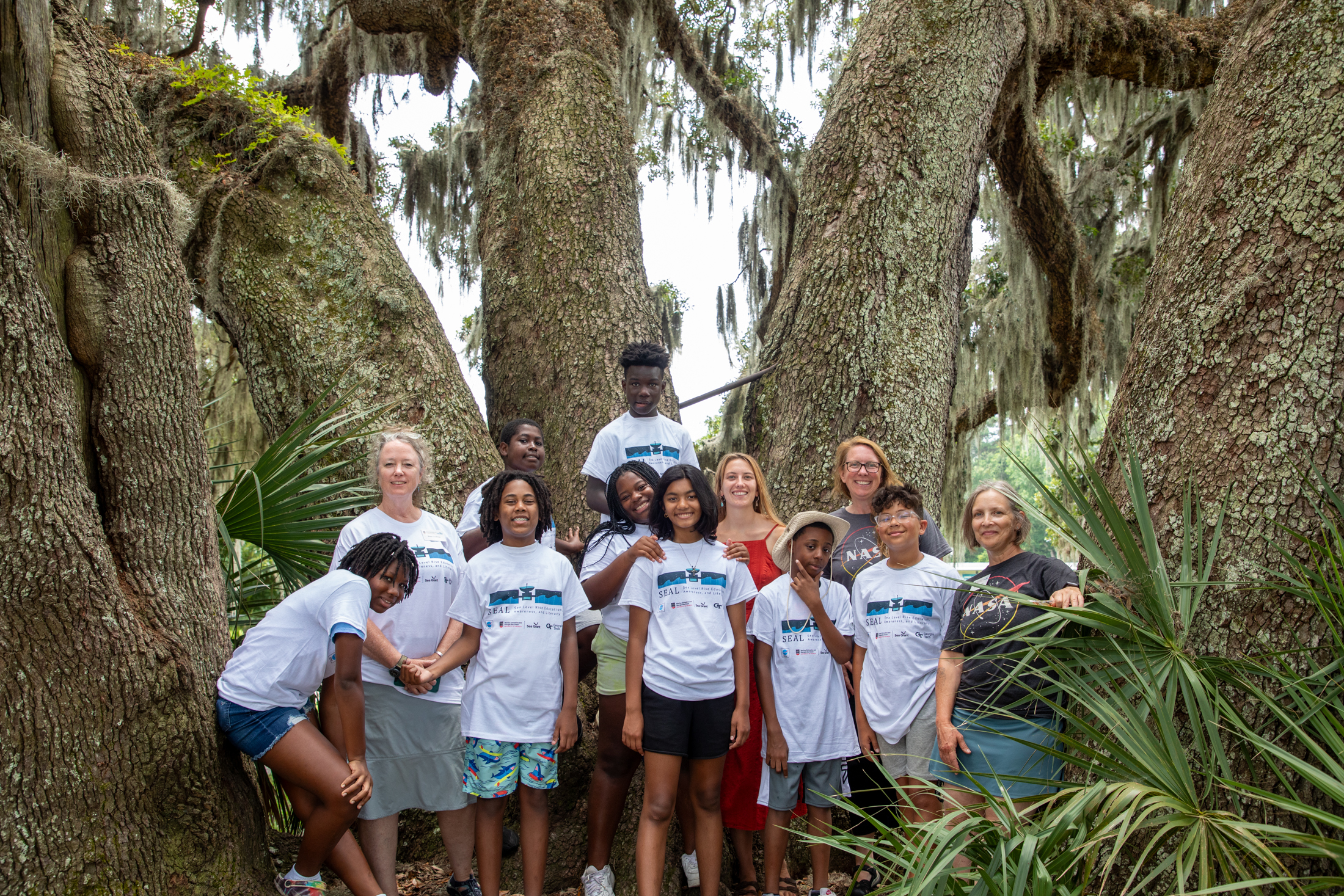 A group of students and two representatives from NASA Science Activation’s Sea Level Education, Awareness, & Literacy (SEAL) team stand in front of some trees as they gather for a group photo.