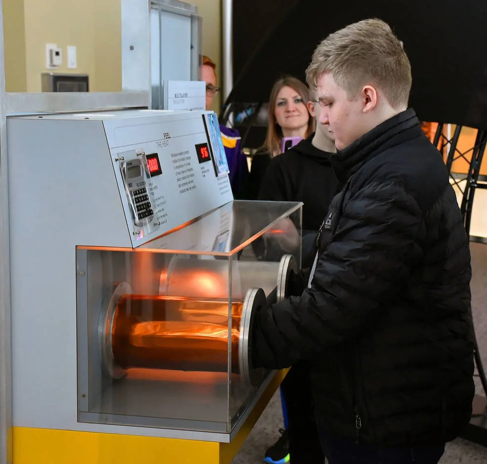 A visitor center guest stands with his hands in two tubes, one with thermal insulation and one without, to understand how temperature is controlled on the spacecraft