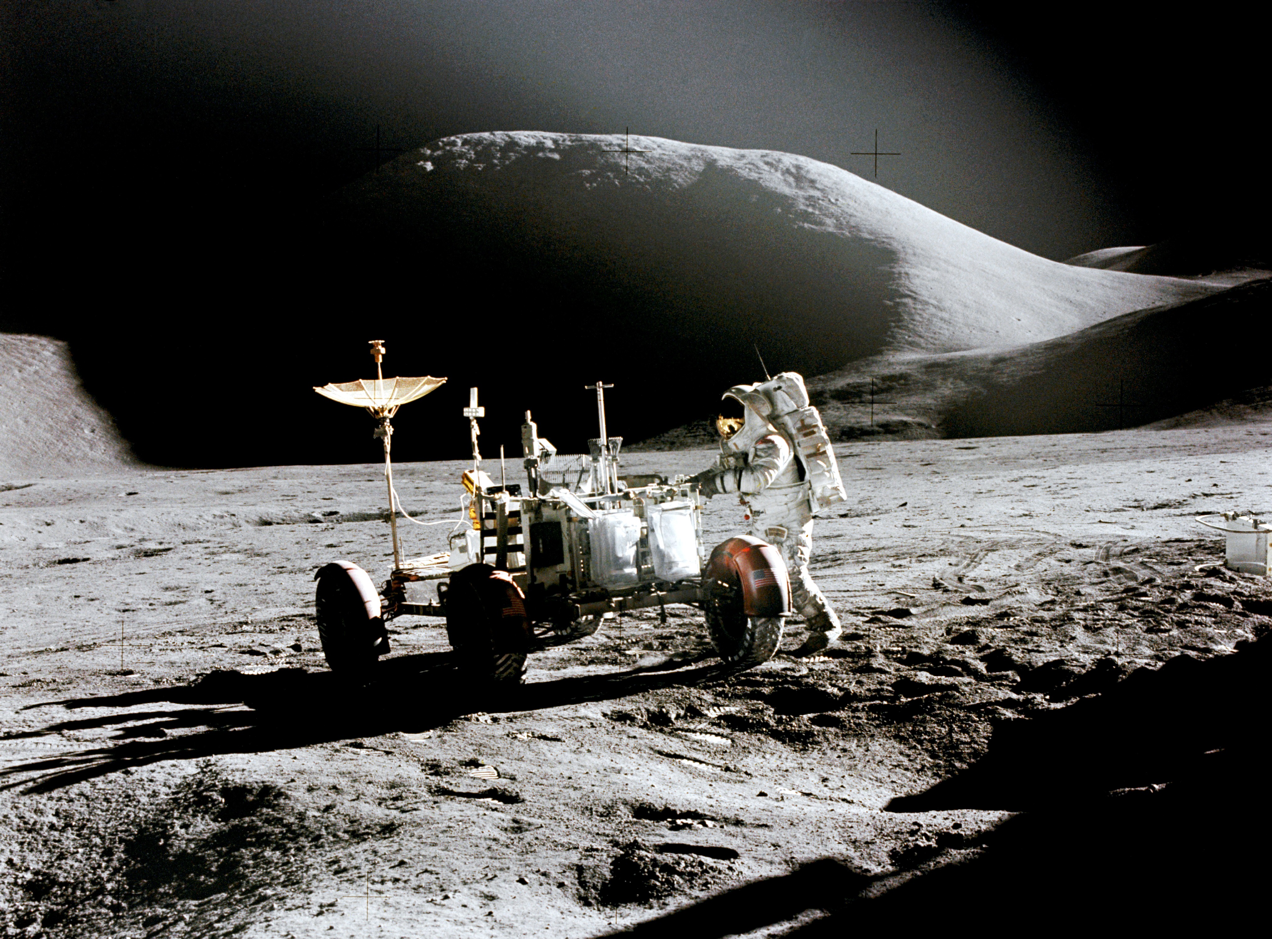 An astronaut is standing on the Moon next to a moon buggy rover. A big hill looms in the background.