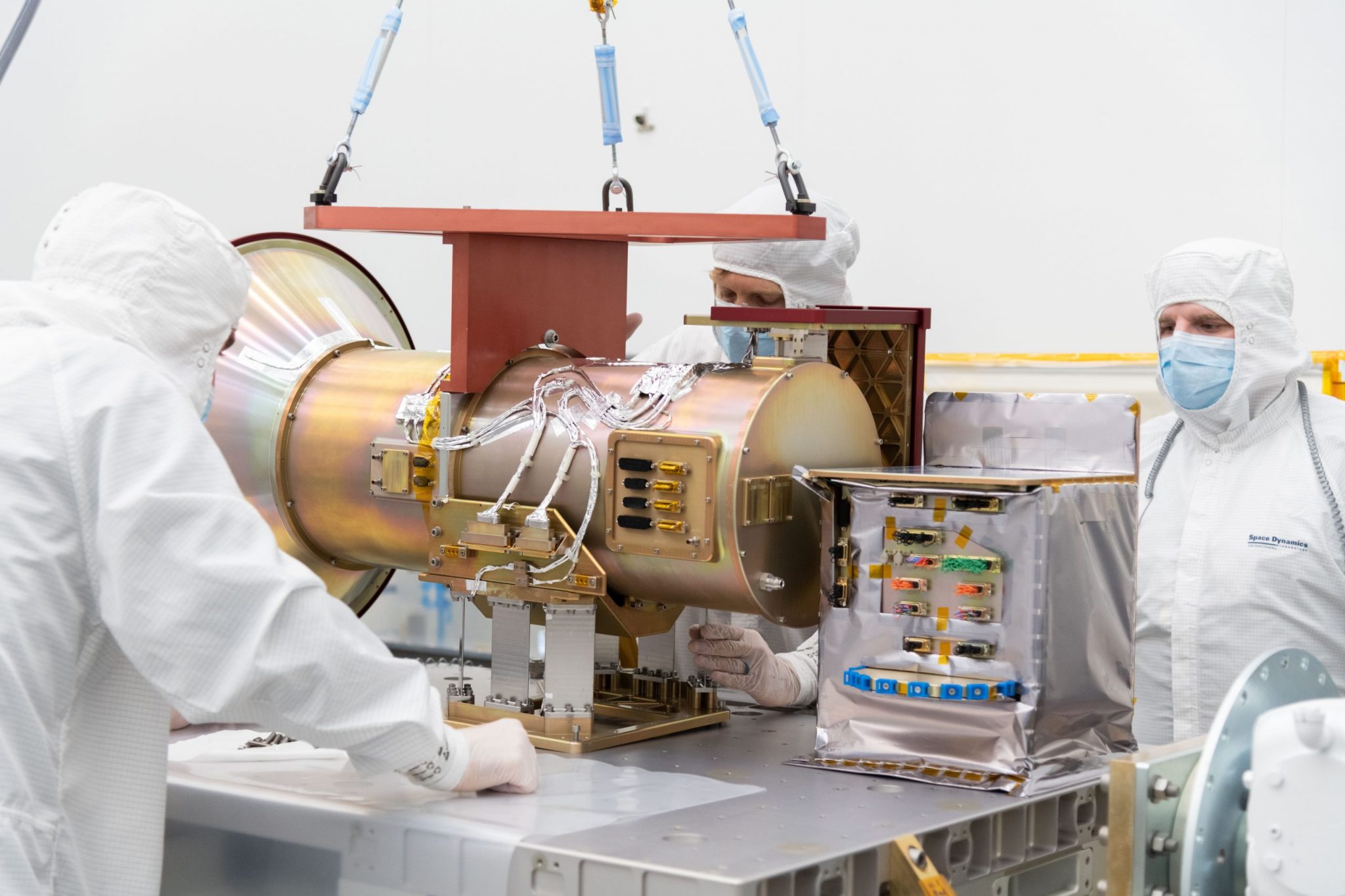 Three scientists in all-white clean room suits stand around a metal table. They are looking at a large, cylindrical metal object with wires coming out of it.