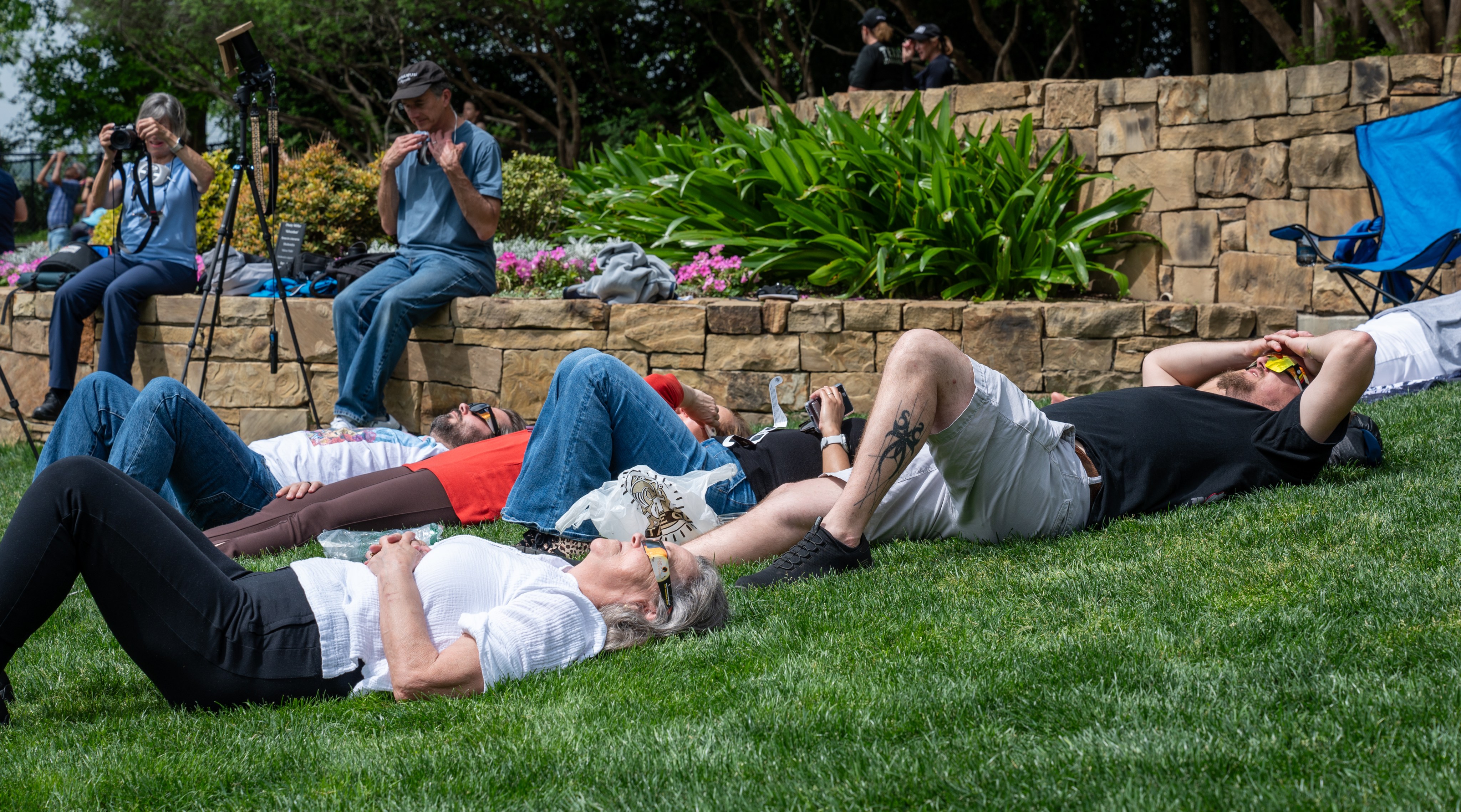 People lay on grass, wearing eclipse glasses, looking up at a solar eclipse.