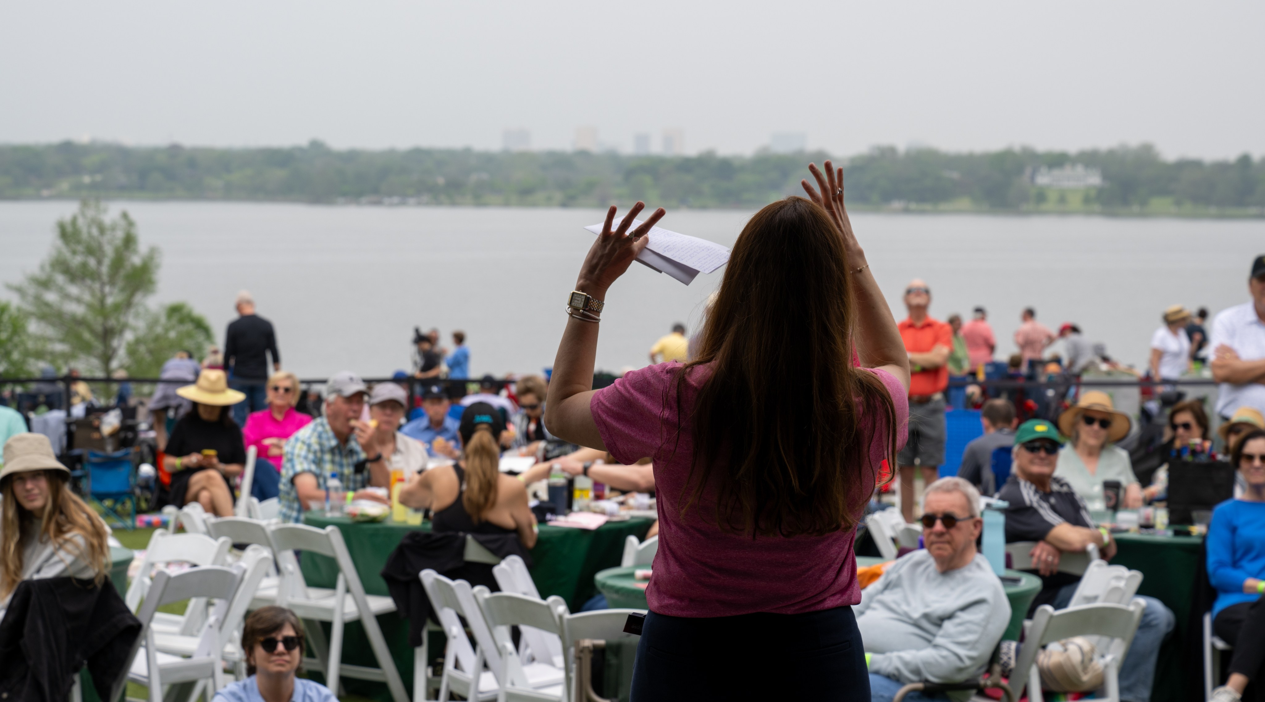 A woman stands with her back to the viewer. She is speaking in front of a crowd, sitting before a body of water.