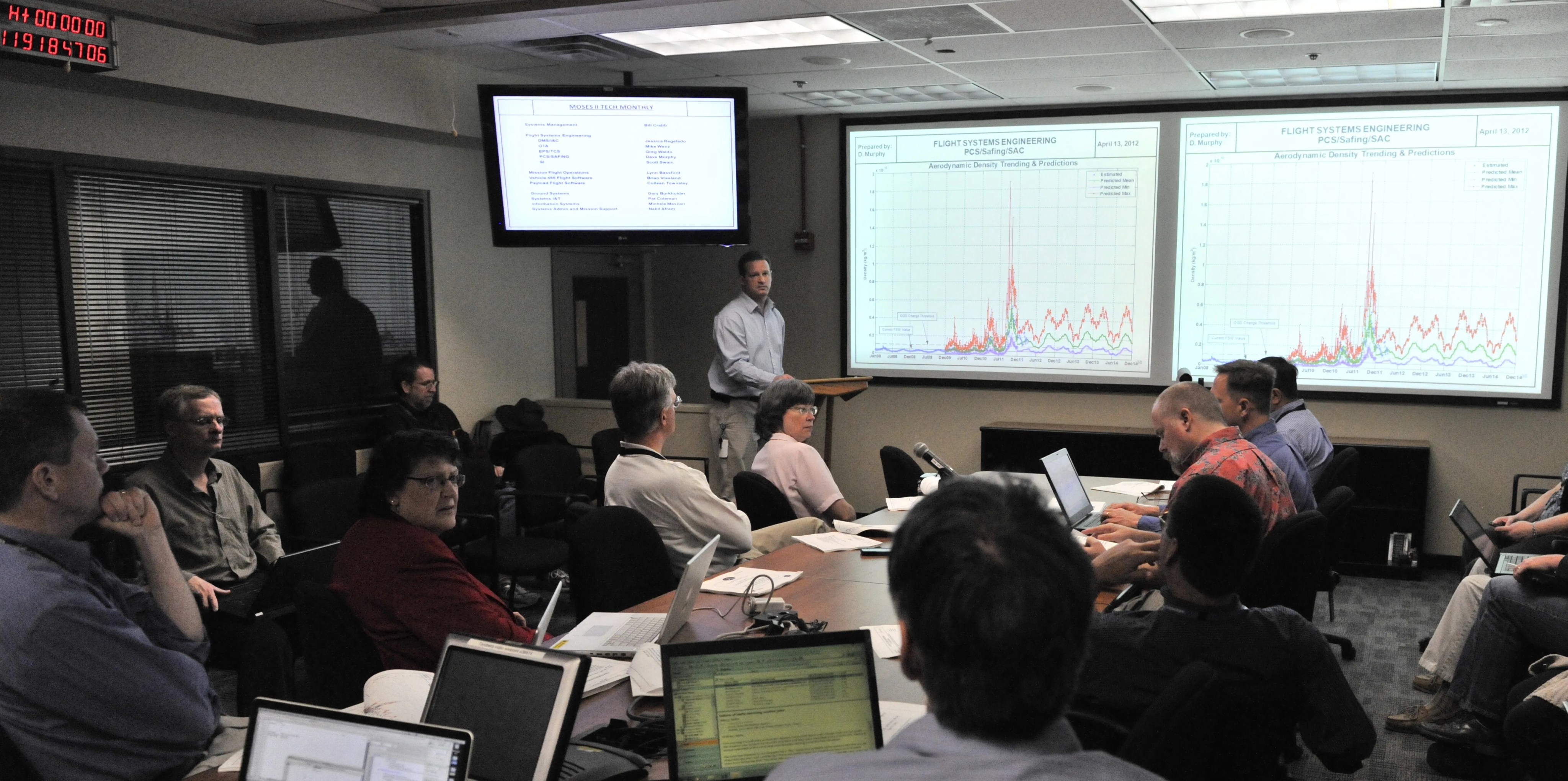 In the background in a conference room, a man presents two charts on the screen at the front of the room for the meeting. In the foreground, men and women sit at a conference table with their laptops, listening to the man present.
