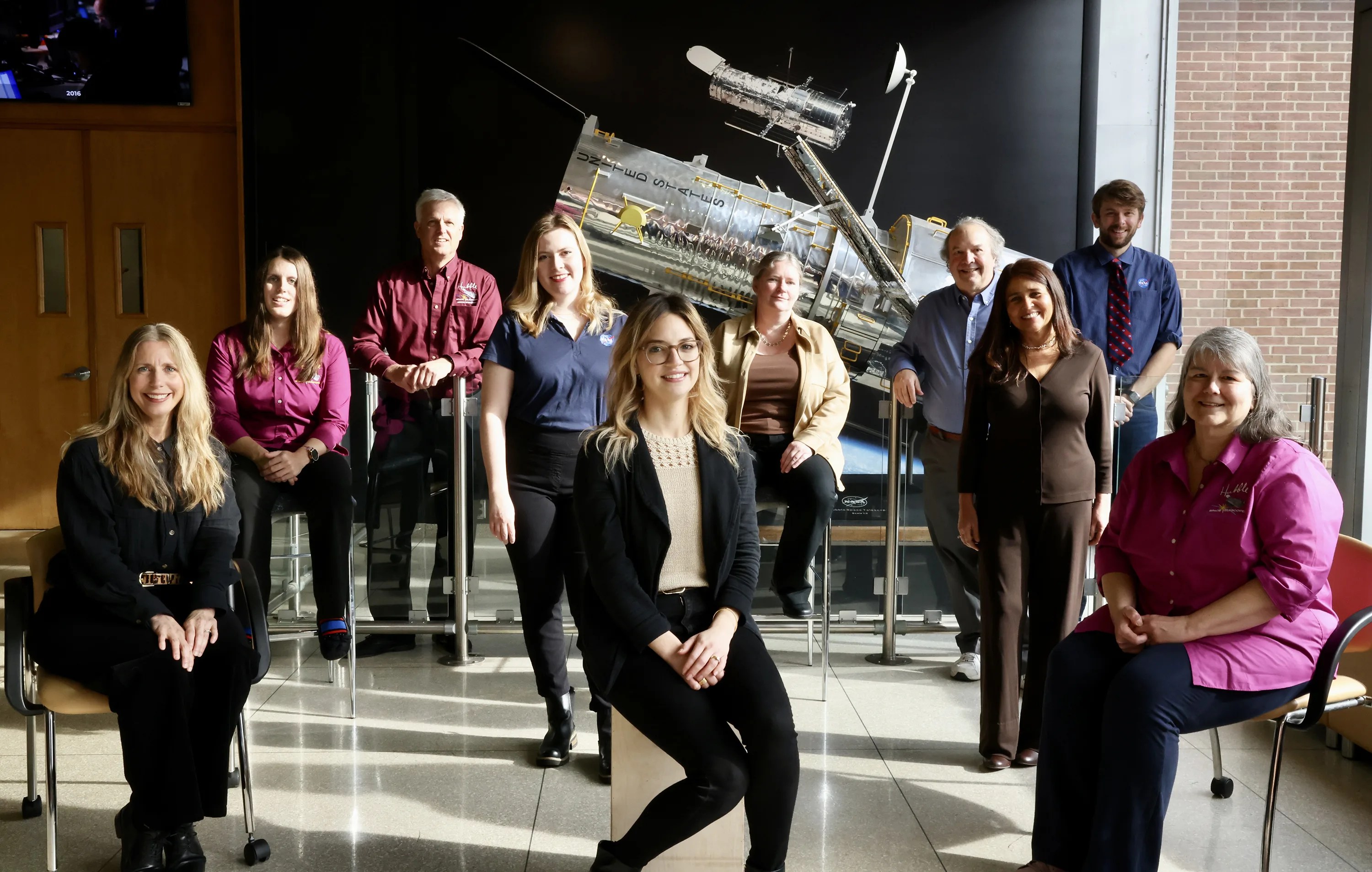 Ten outreach team members standing and sitting posing in front of a scaled model of the Hubble Space Telescope.