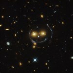 Two bright orange galaxies seem to form eyes, and streaks of curved, bent and magnified light form the circle of a face and a happy smile. A smaller bright galaxy serves as the nose in this smiley face. Other galaxies dot the picture.