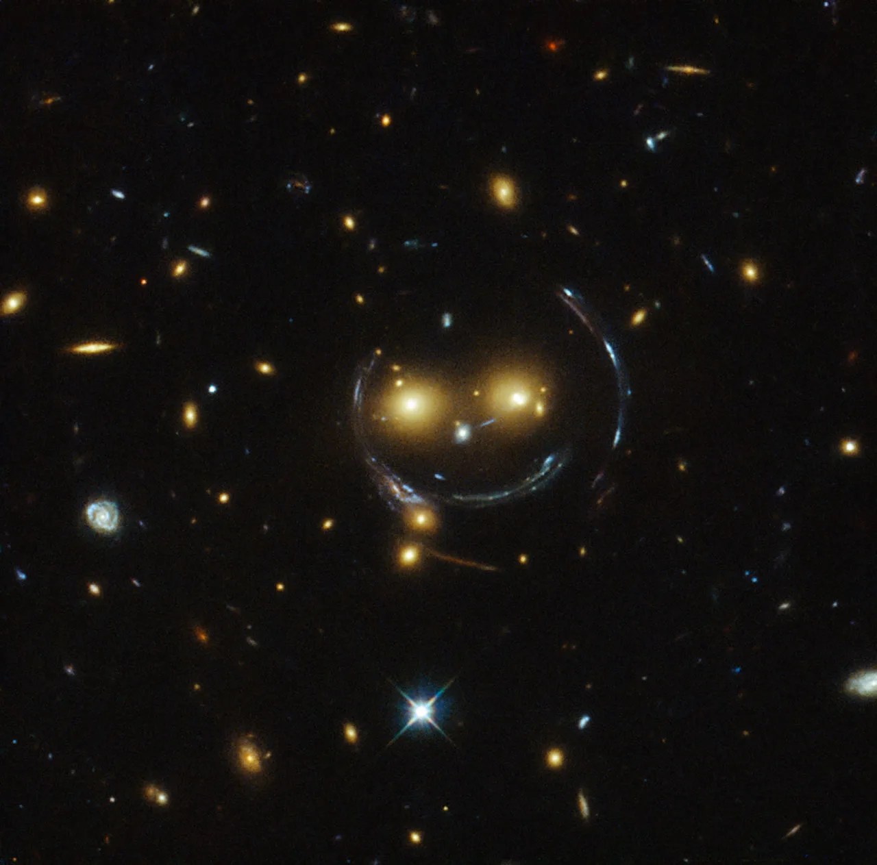 Two bright orange galaxies seem to form eyes, and streaks of curved, bent and magnified light form the circle of a face and a happy smile. A smaller bright galaxy serves as the nose in this smiley face. Other galaxies dot the picture.