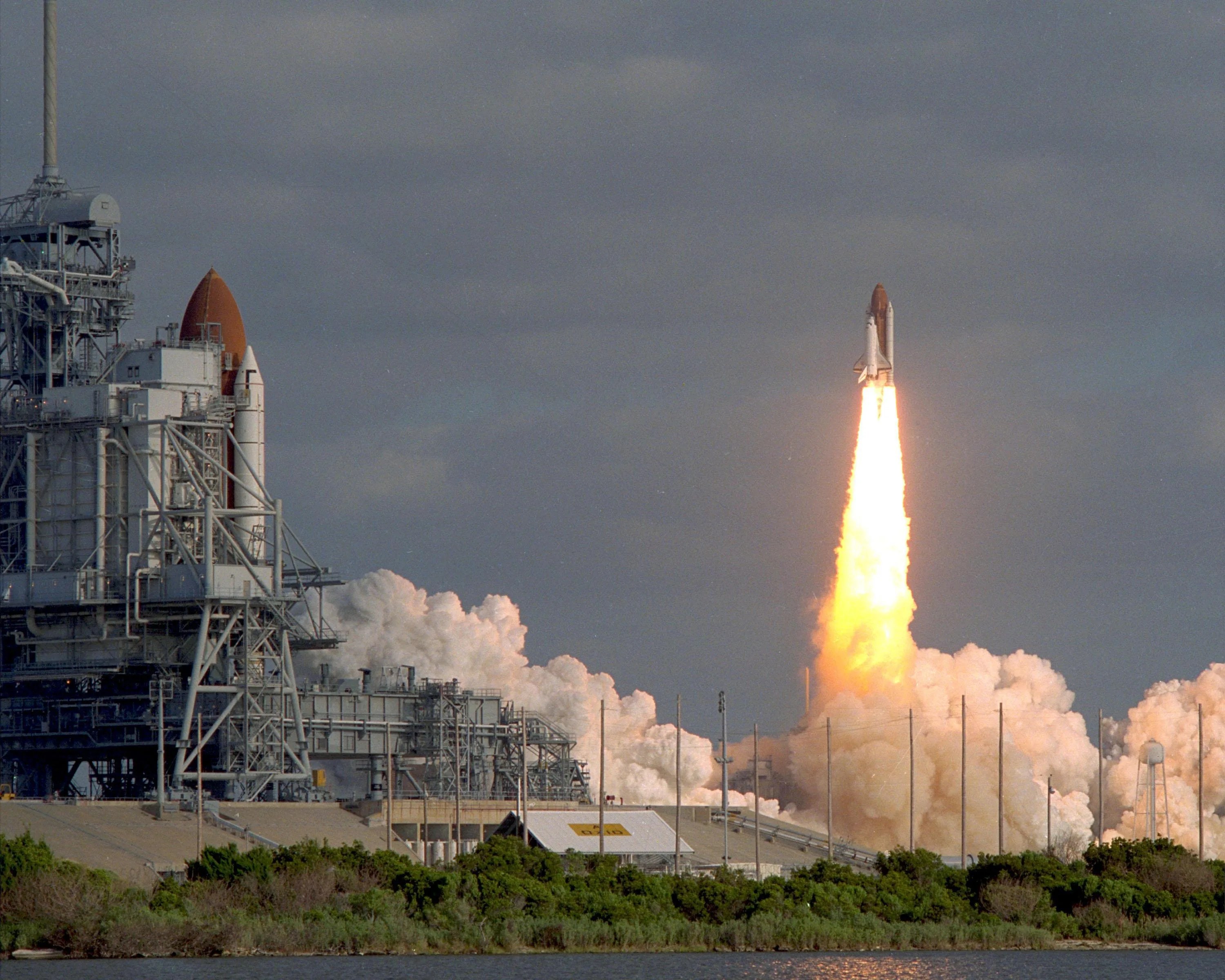 In the distance, shuttle discovery launches in daytime, with shuttle columbia on launch pad visible at far left of image