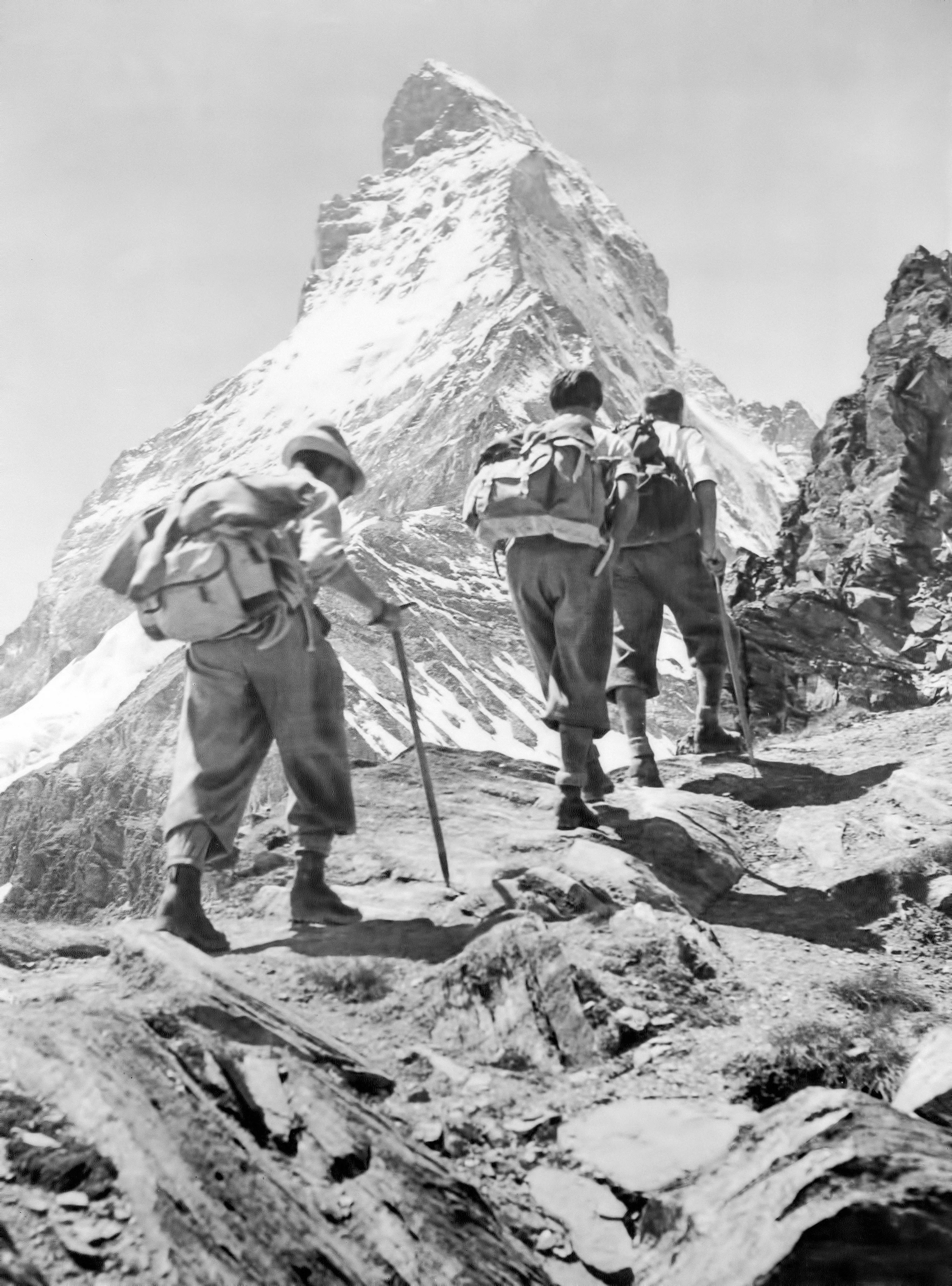Three men hike up a rocky terrain with snow covered mountains in the background.