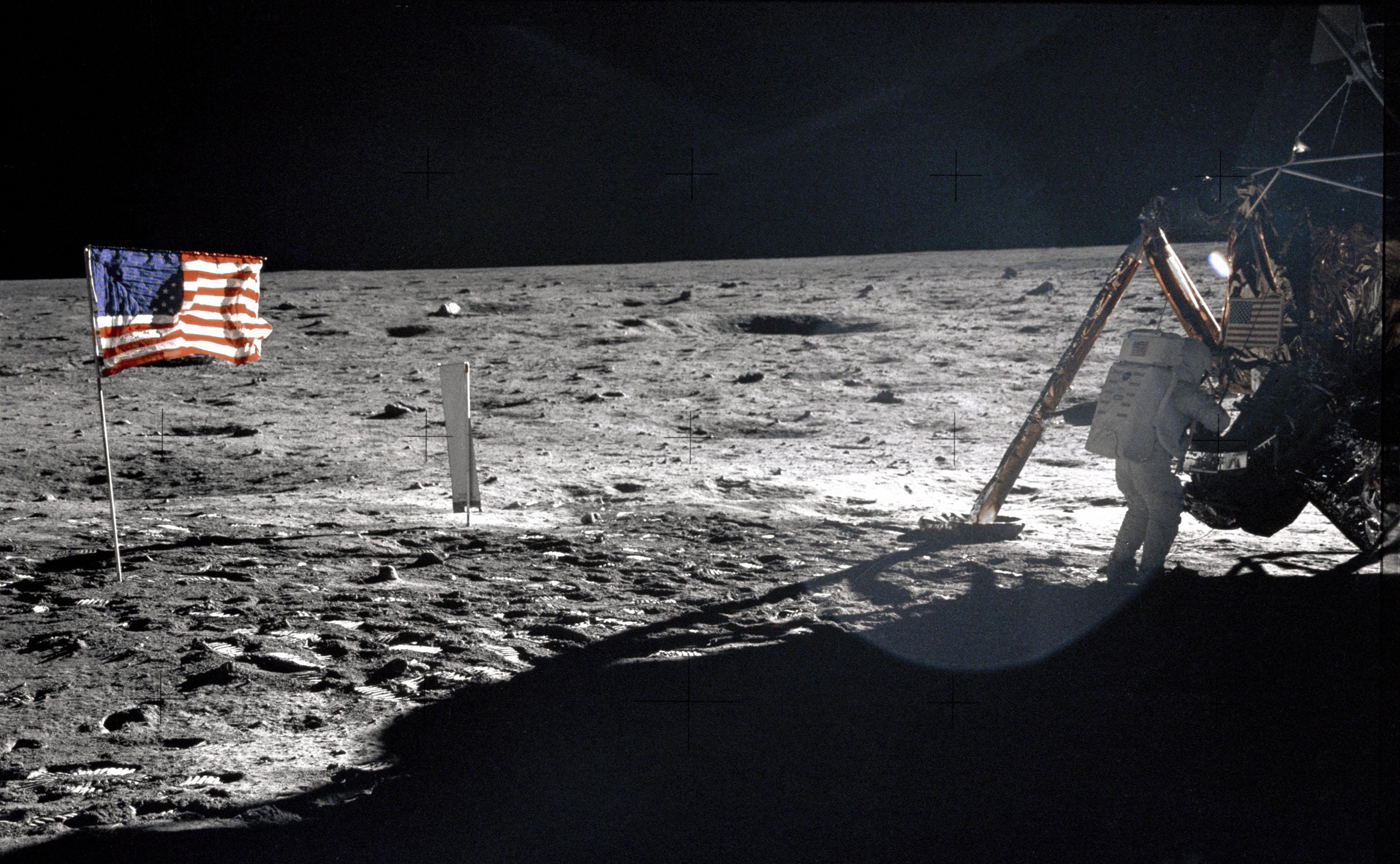 To the far right of this image, an astronaut in a white spacesuit has his back to the camera while he works near the lunar lander. To the far left is an American flag on a pole in the lunar soil.