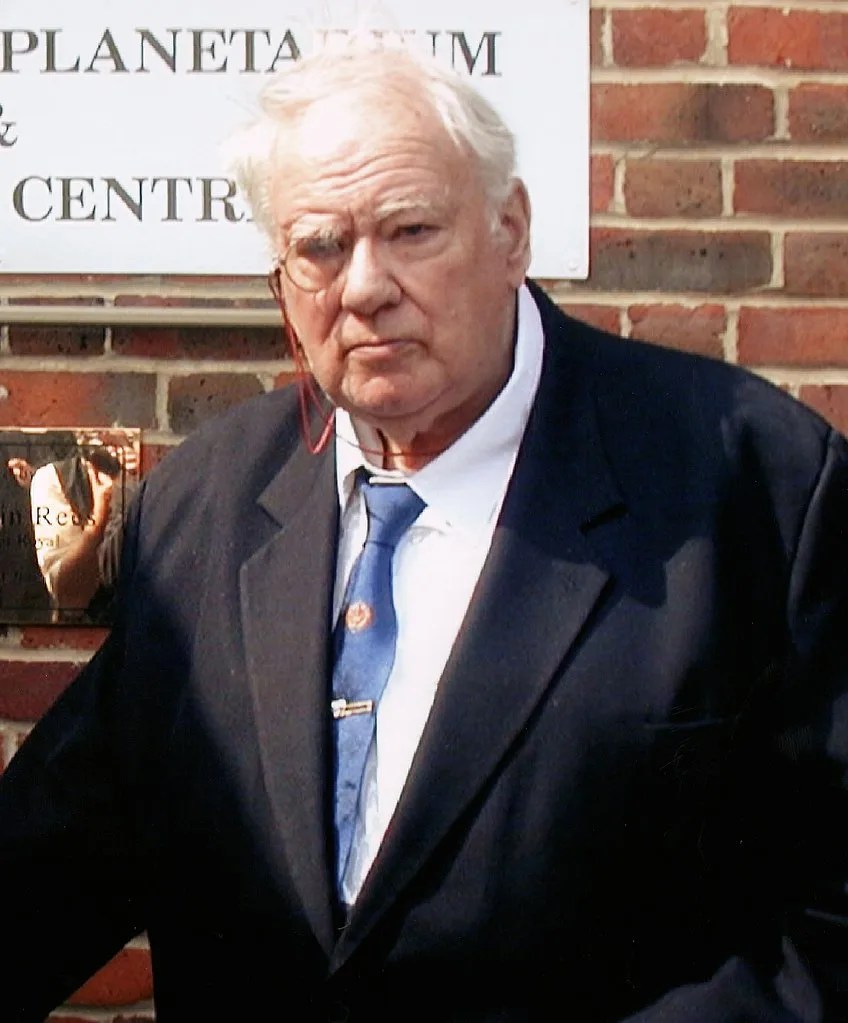 headshot of an older man with gray hair and a monocle on his right eye standing in front of a brick wall