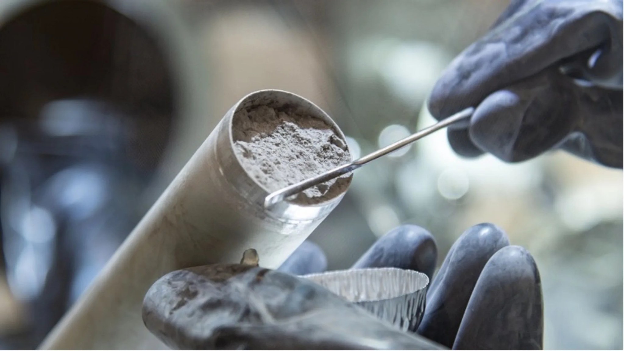 The image consists of a hollow silver colored cylinder that is filled with a gray lunar soil. A scientist wearing a pair of dark gloves is collecting a tiny sample of the lunar soil from the cylinder using a long, thin wand.