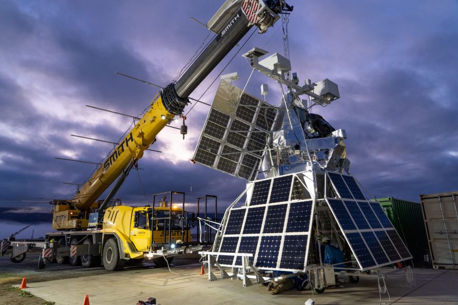 he SuperBIT payload hangs from a yellow crane vehicle. The payload is made up of multiple solar panels, a bar across the top with various instrumentation, and many electronic components make up the main body of the payload. The sky in the background is dark and heavily clouded over.