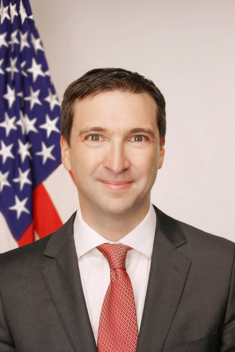 Portrait photo of Dr. Dan Evans wearing a dark suit and red tie in front of the American flag.