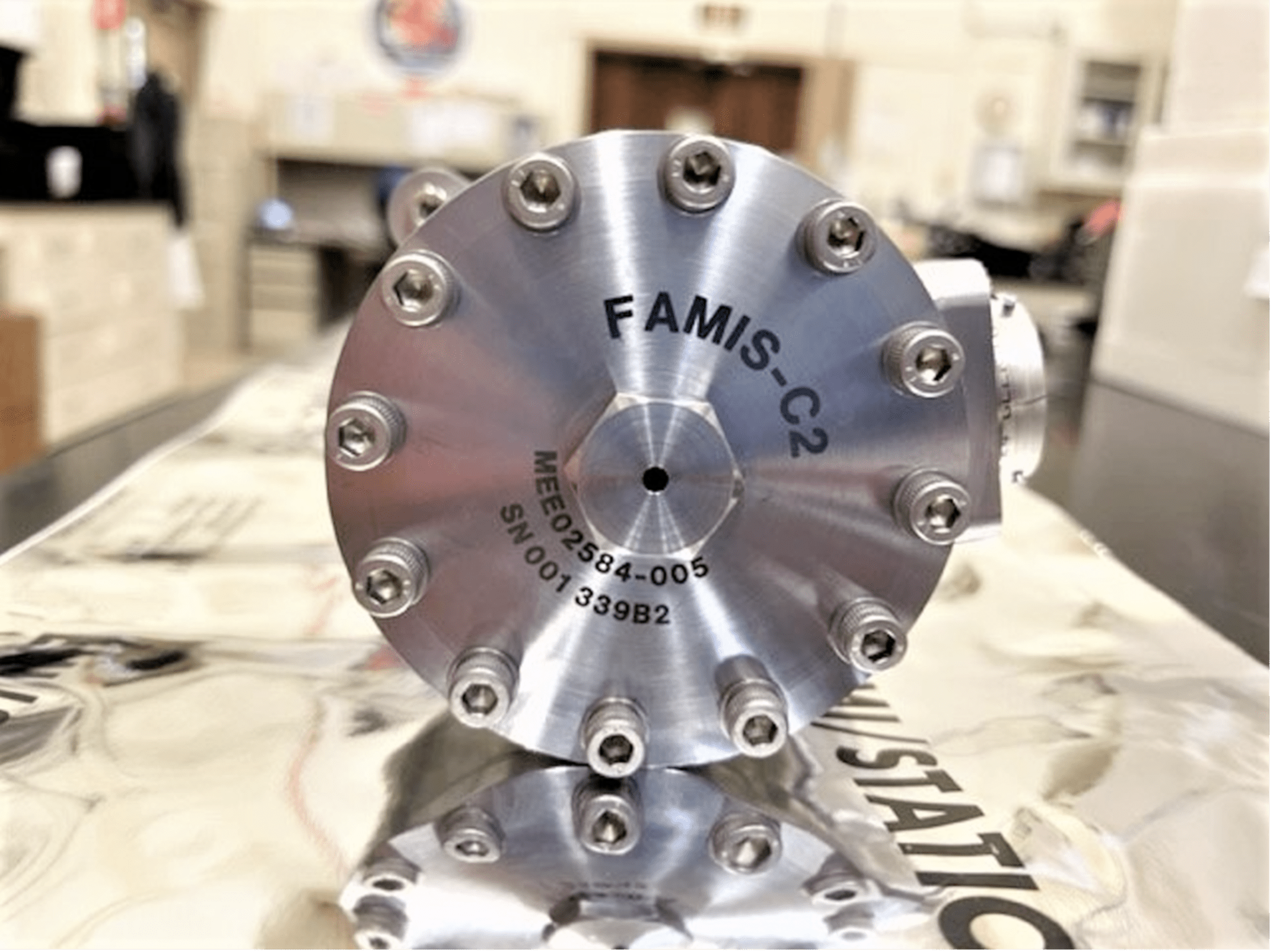 Image shows the circular side of a metallic Sample Cartridge Assembly (SCA). Twelve bolts line the circumference of the device