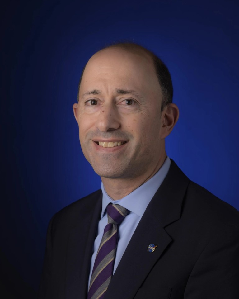 Portrait photo of a smiling man wearing a dark suit jacket and tie and sitting in front of a dark blue background that fades to black.