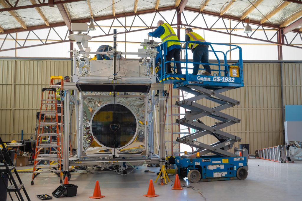 Two NASA technicians wearing reflective vests are atop a scissor-lift and reaching over the top of the Extreme Universe Space Observatory-2 payload, which is cubed-shape with a circular lens on one surface.