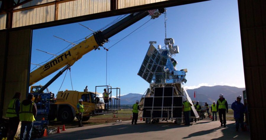 The SuperBIT payload hangs from a yellow crane vehicle. The payload is made up of multiple solar panels, a bar across the top with various instrumentation, and many electronic components make up the main body of the payload. The sky in the background clear and virtually cloudless.