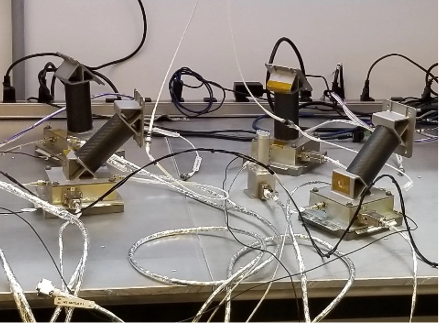 Four sensors sit on a table connected with a series of silver-colored wires.  Each sensor consists of a silver base plate with a black cylinder attached at a 45-degree angle. At the end of the cylinder sits a silver flat plate, which angle varies from sensor to sensor.