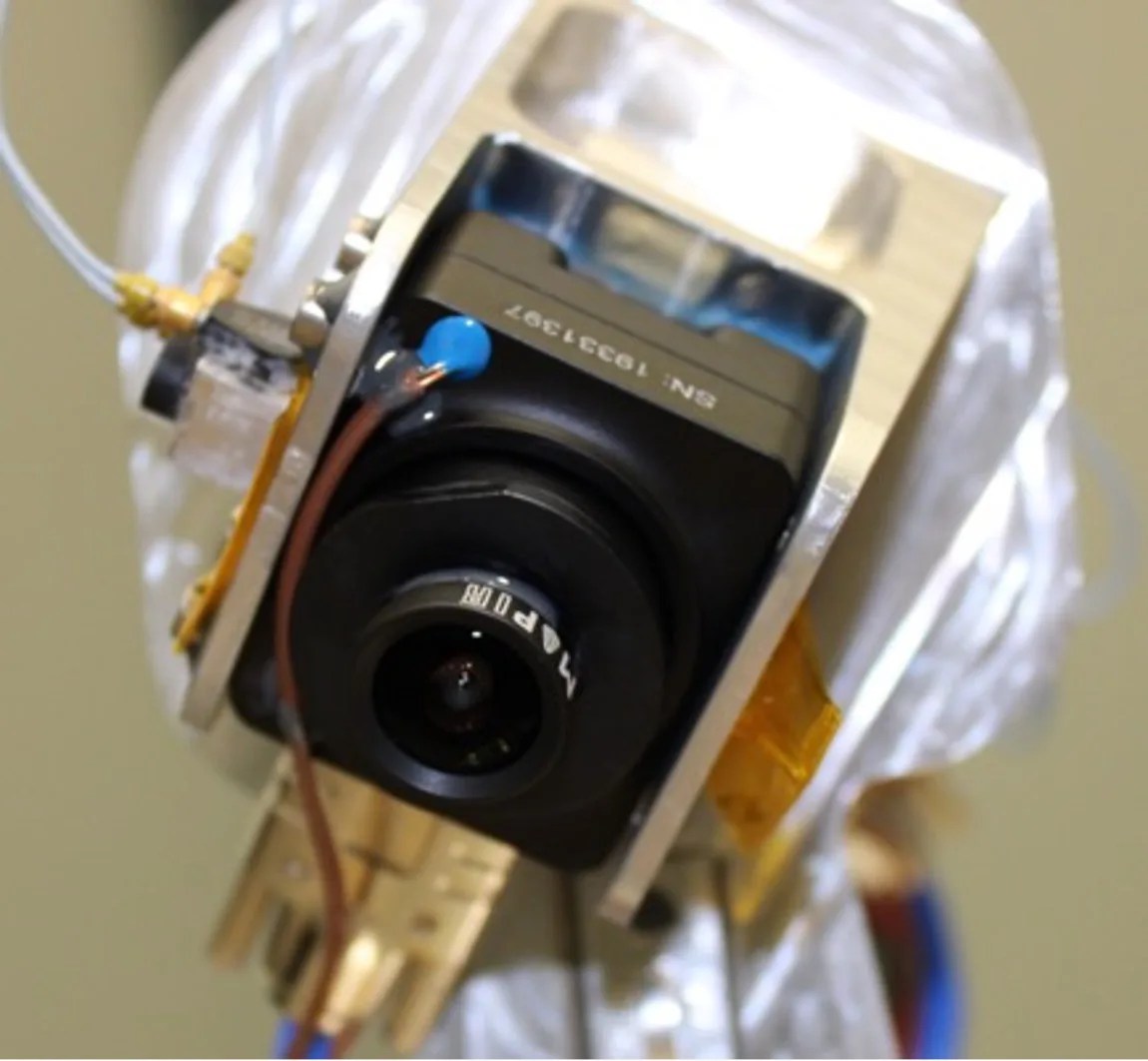 One of the SCALPSS cameras is attached securely to a metal receptacle. The section directly touching the camera is golden and the larger mount behind is silver. Wiring connects the camera to items outside the frame: a silver pair of wires is attached at the top and red wire is attached to the front with blue adhesive.