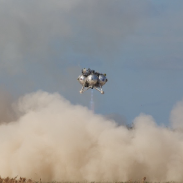 SCALPSS is hovering near the ground above a sizable dust cloud caused by lander plume. The shape of the structure consists of four silver spheres clustered adjacent to each other, with each sphere containing a camera. Two pairs of adjoined legs extend down from the bottom. In the background, a blue-sky is visible.