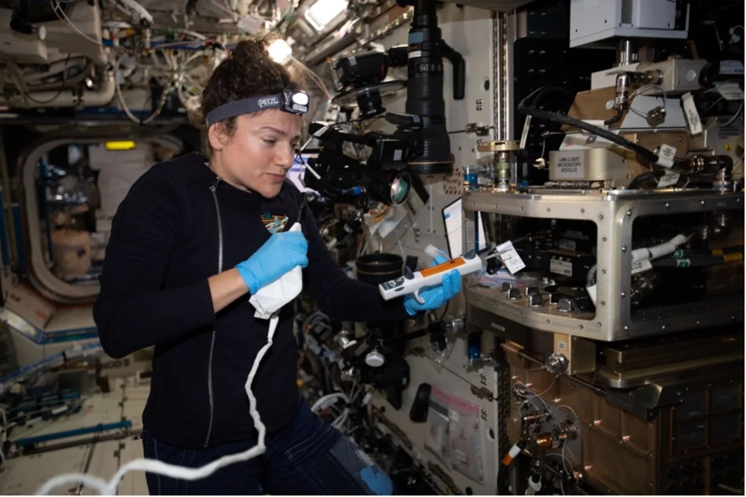 Astronaut wearing blue gloves looking at a white and orange device in her left hand while holding onto a white clothed device attached to a tether in her right hand.