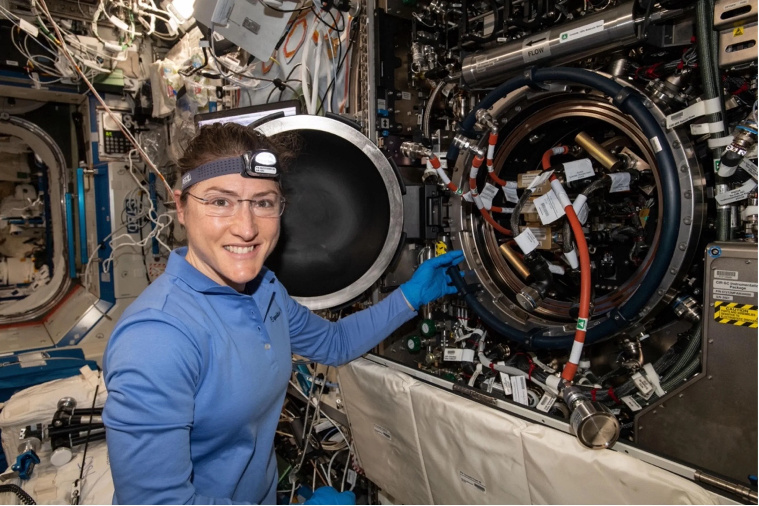 Female astronaut next to an open circular area onboard the International Space Station with red tubes and other mechanics inside.