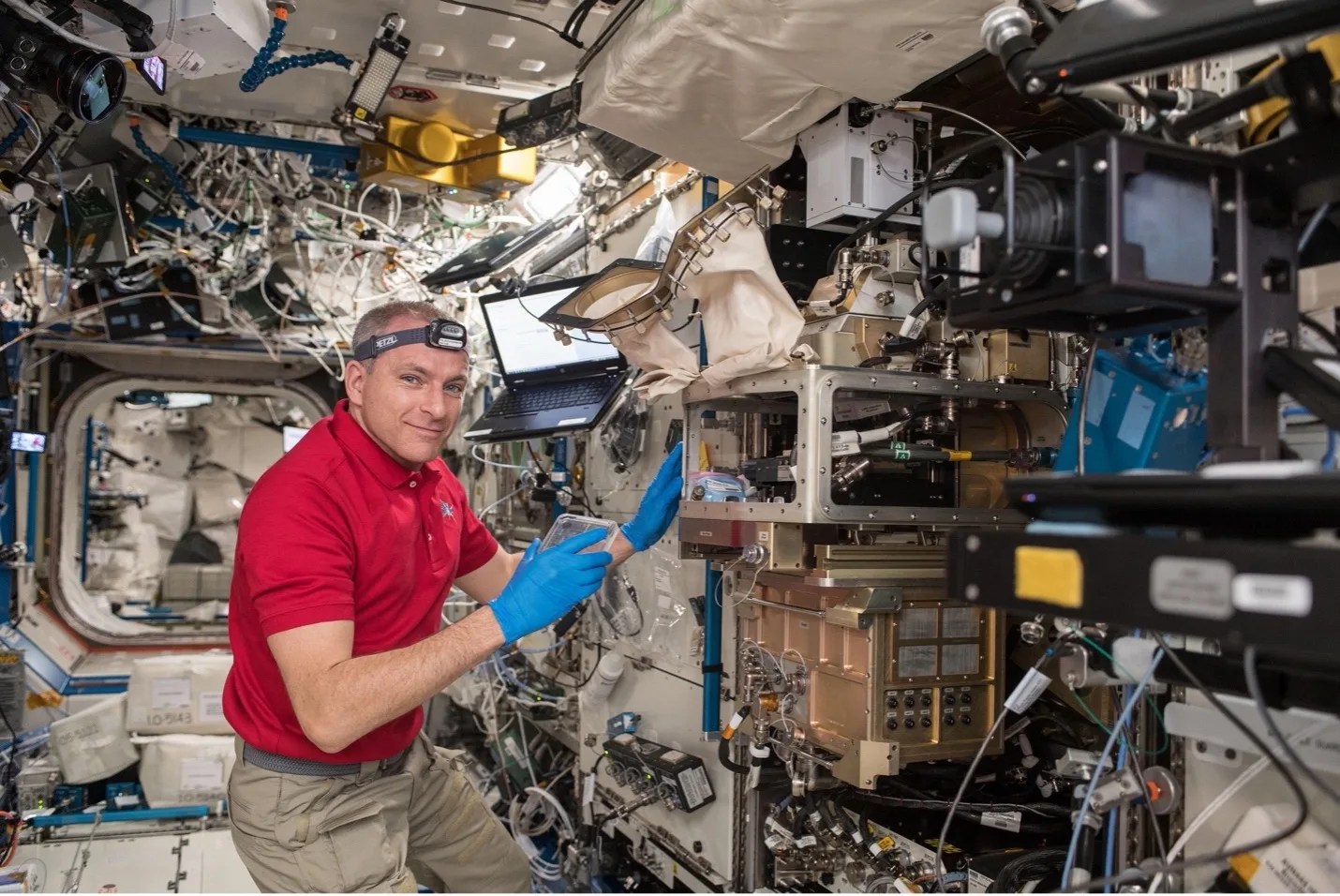 Astronaut surrounded by wires and mechanical devices within the space station. He’s wearing blue latex gloves while handling a transparent flat device in his right hand and holding onto a rectangular metal frame attached to the wall.