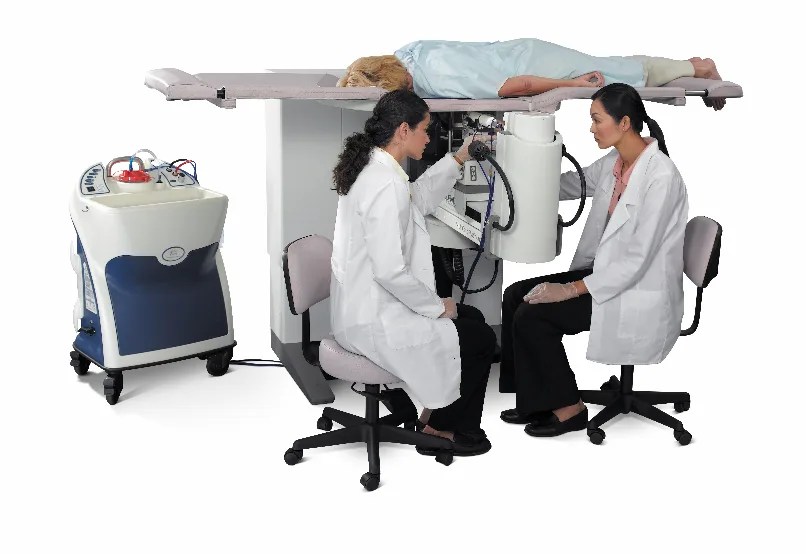 Two medical workers in white coats work at an imaging machine. A patient in a hospital gown lies on a platform on top of the machine.