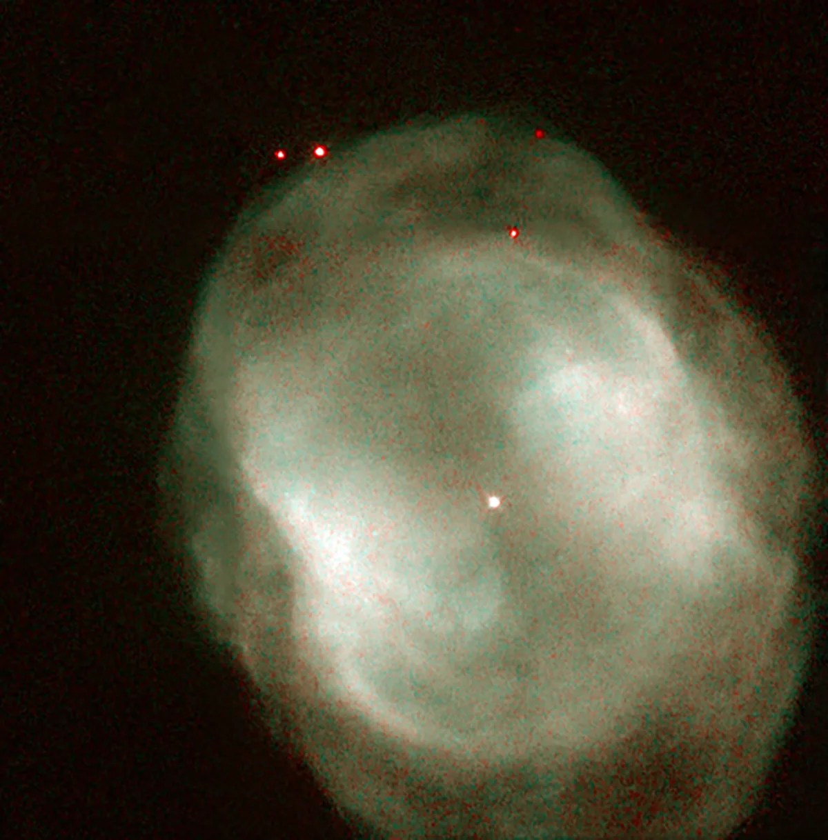 A grayish oval nebula with a star at its center. Four red stars are located near the top of the nebula.