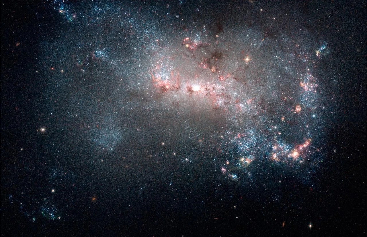 Large grouping of stars and bluish dust and gas. Near the center the stars create a bright white light. Galaxy is a amorphous blob.