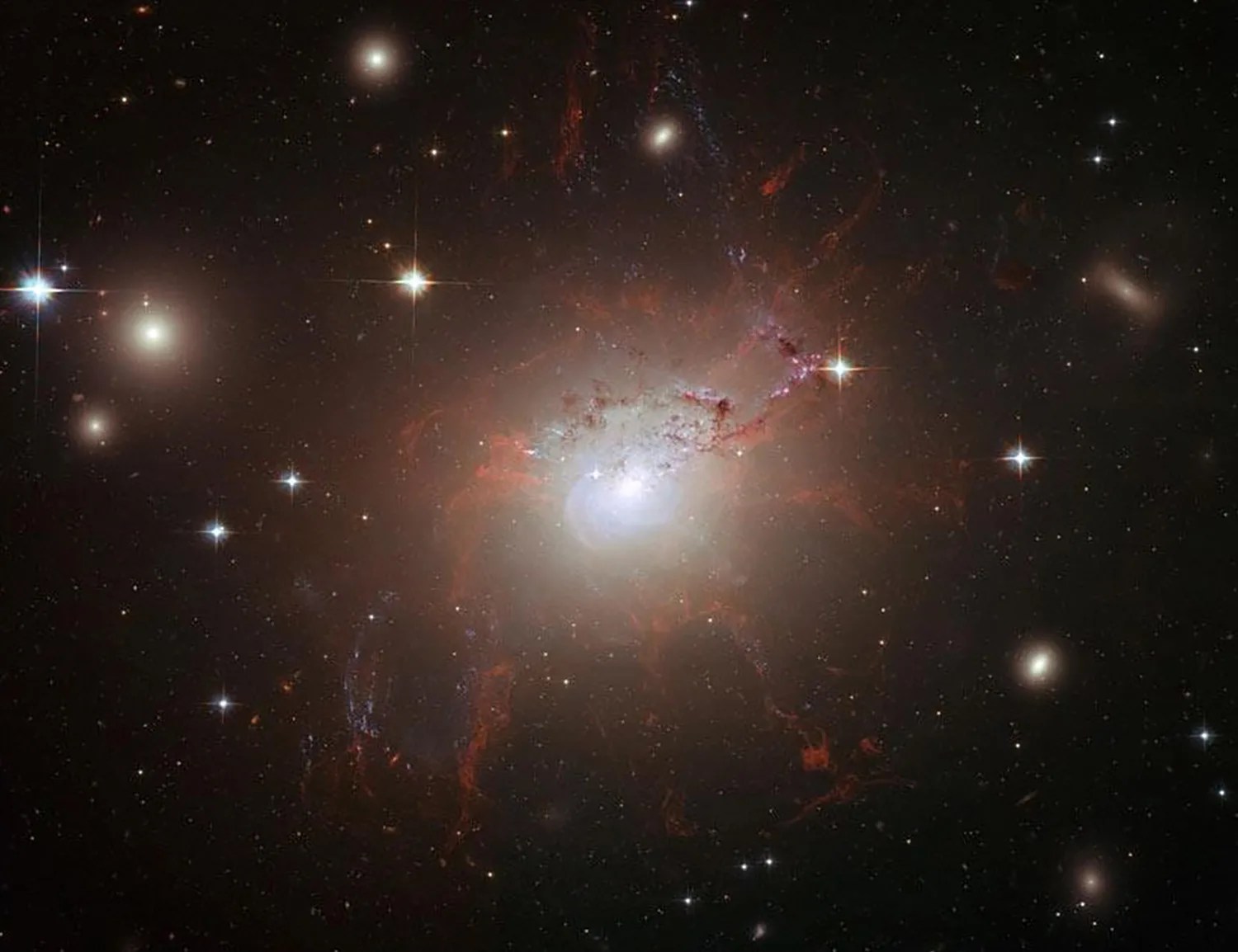 Large glob of stars surrounded by dark rusty red gas and dust. Huge bright stars surround the bright galaxy in the center.