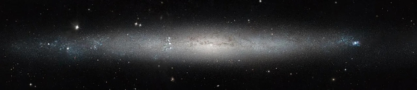 Long thin galaxy view, nearly from the side but tilted a bit toward the viewer. Lots of white stars throughout, but they get thicker and brighter near the center.