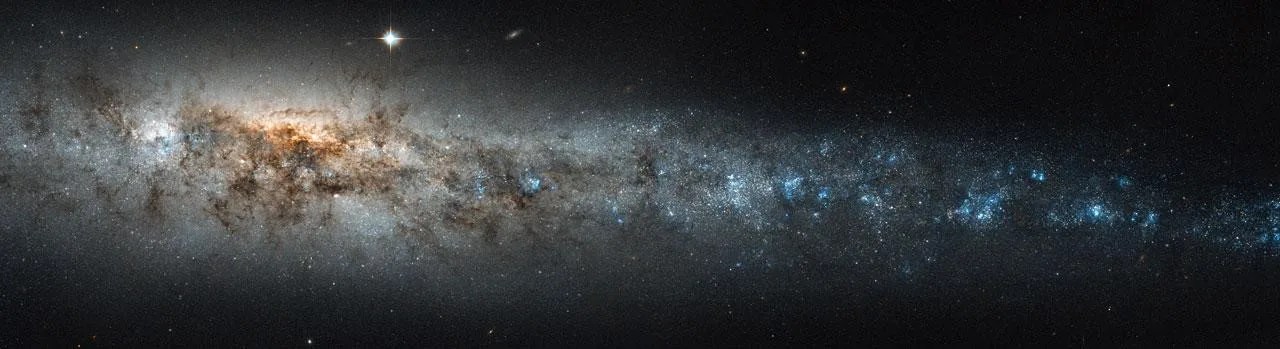 Long thin side view of a galaxy. On the left side, a larger part of it, closer to the central core. Lots of gas and dust surrounding countless stars. The stars softly stretch out to the right of the image.