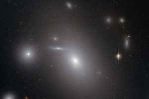 Large, egg shaped, white galaxy in the center, surrounded by stars and dust, other galaxies behind the center one peppered throughout.