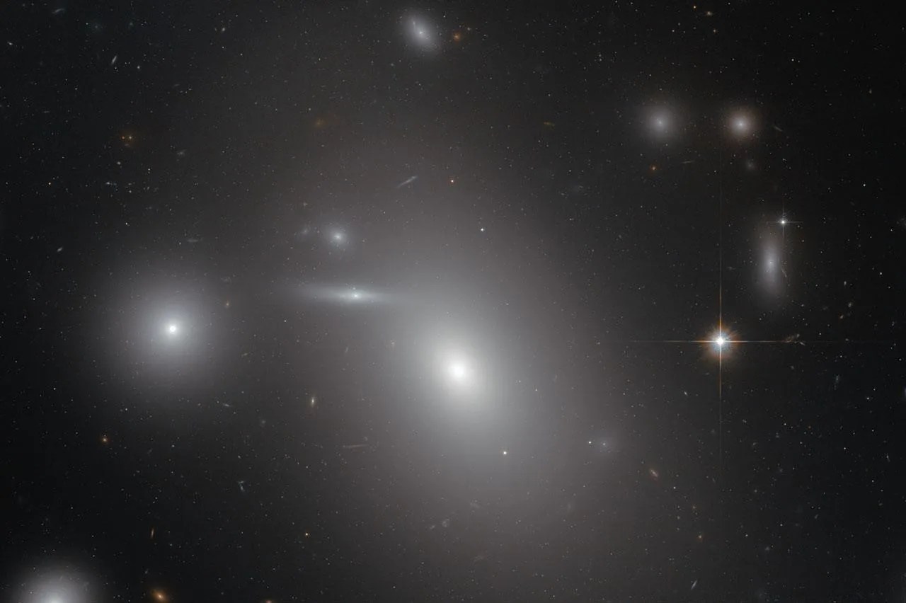 Large, egg shaped, white galaxy in the center, surrounded by stars and dust, other galaxies behind the center one peppered throughout.