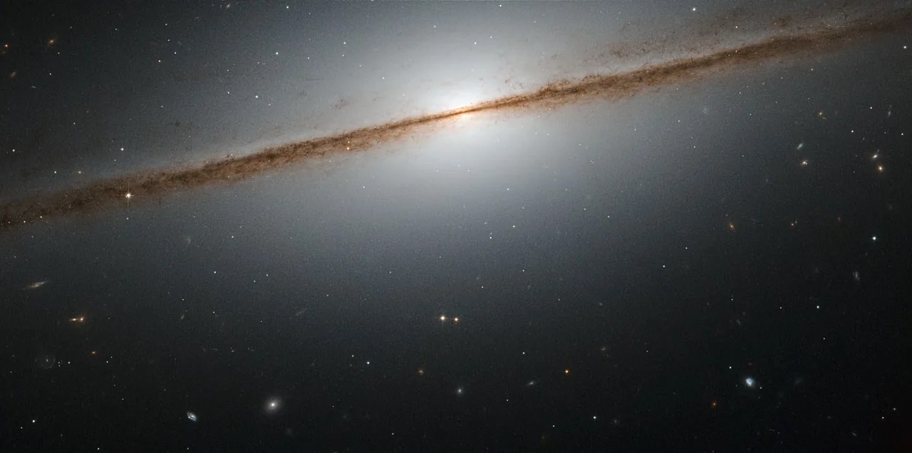 The pinkish-brown disk of a spiral galaxy, viewed from head-on and looking like a straight line, stretches across the picture. The center of the galaxy is glowing white and looks spherical, and a white glow radiates outward from it, encompassing the whole galactic disk.