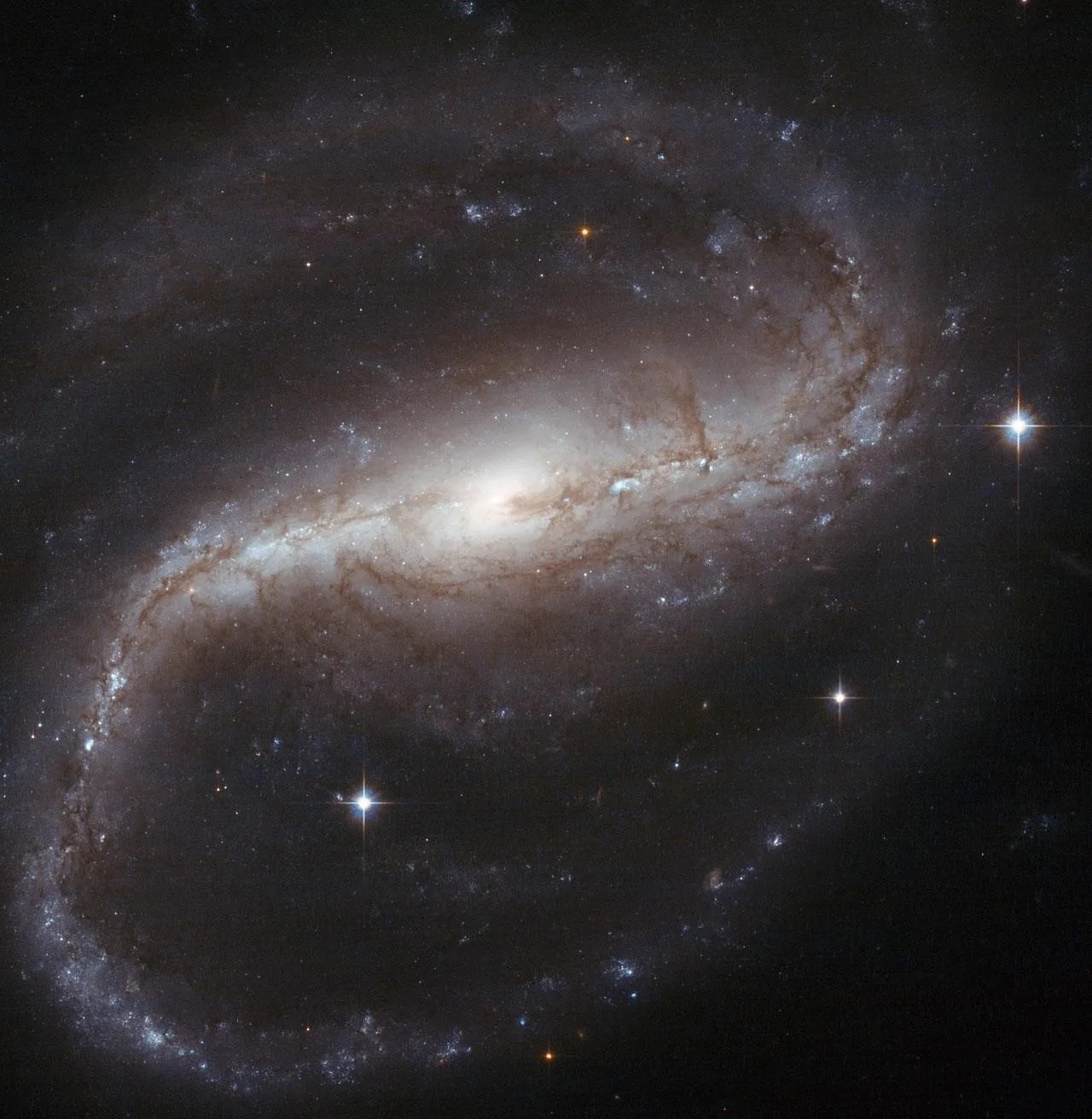 A "barred spiral" galaxy is shaped like a backward S, with its arms of gas and dust made up of whites, pinks, red-browns, and pale blues. The center of the galaxy (the middle of the S) is a glowing white sphere.