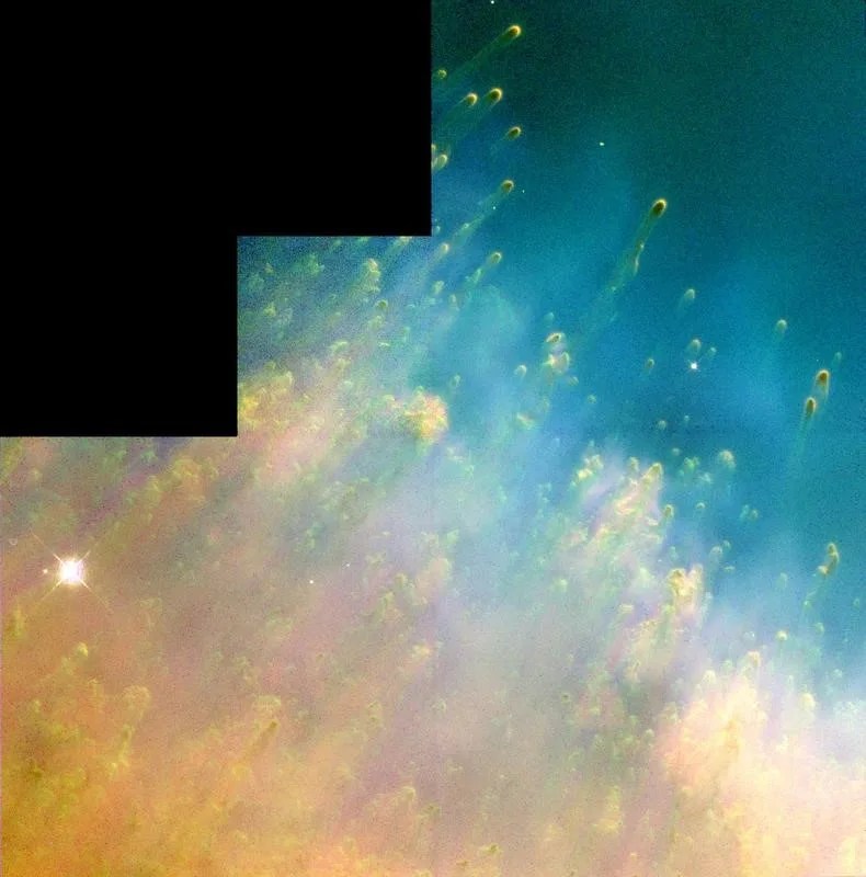 The lower left corner of gas and dust is a bright orangish-yellow. Streams of gas resembling tadpoles flow up and to the right from this cloud, which fades into a pink-yellow, then to a green-blue, and then into blue.