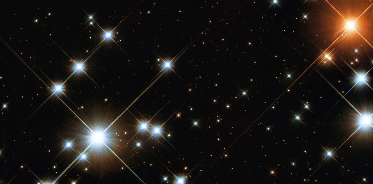 Several bright white stars with x-shaped spikes of light are located to the left of the image. A few more, inclucing some reddish stars, are at the edge of the image to the right. Smaller scattered stars are in the background against black space.
