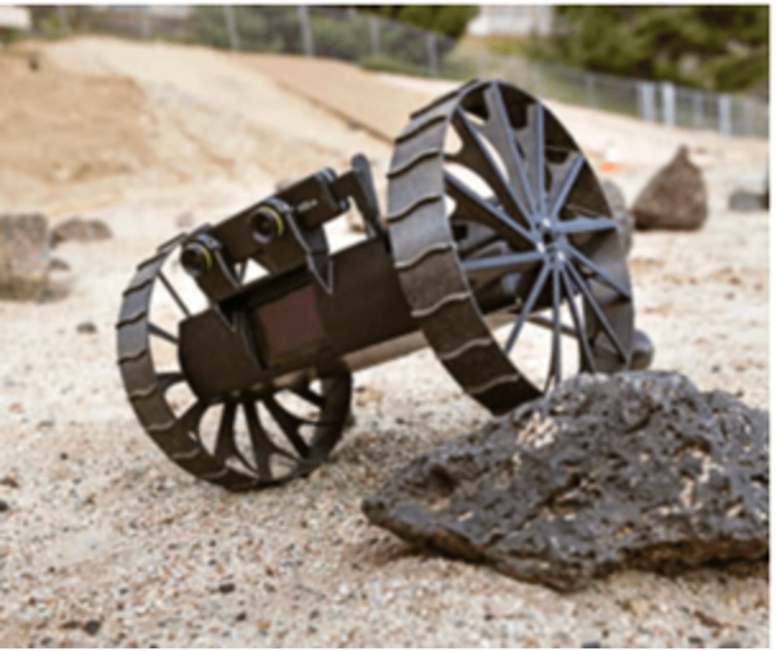 A small, motorized robot rolls on the surface of a sandy area. The robot has two large wheels and the one on the right is driving over a large black wheel. The overall color of the rover is a dark black while the sand contrasts as a tan color.