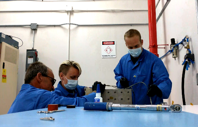 Two men and one woman in blue lab jackets work on HaloSat hardware on a table.