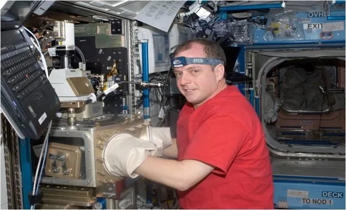 Astronaut with his hand inside large, white latex gloves attached to a gold, mechanical device.