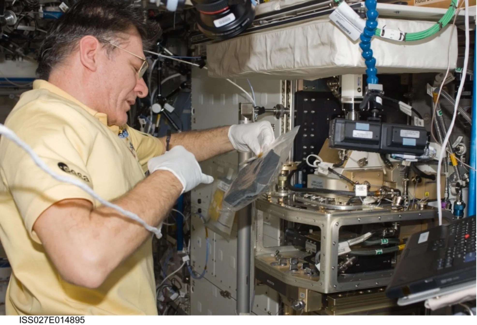 Astronaut wearing white latex gloves holds a small microphone attached to a white wire. As he speaks into the microphone using his right hand, he holds a black square device within a transparent bag with the other.