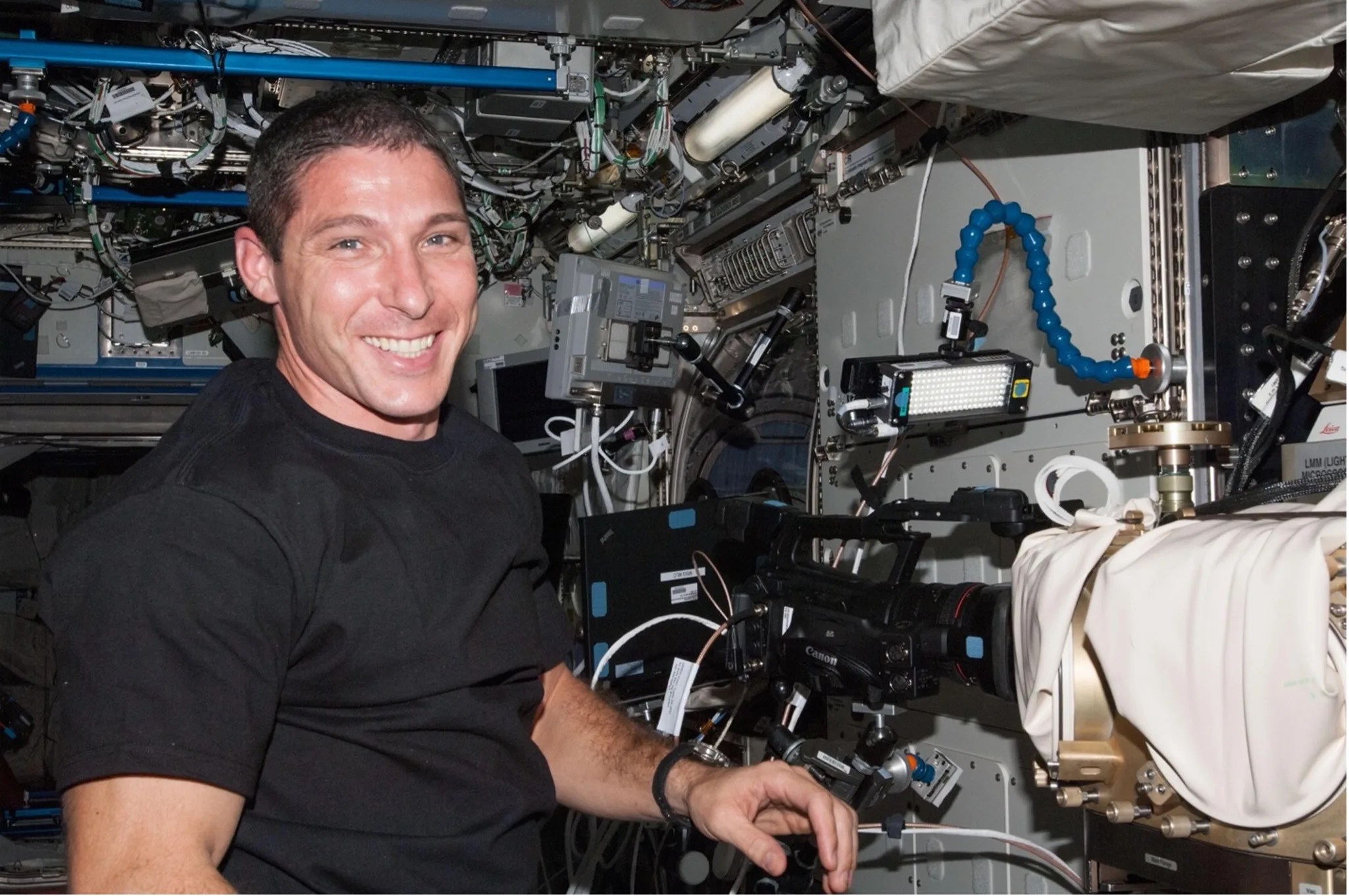 Astronaut within the space station stands next to a large black video camera. Surrounding him are wires and mechanical devices including a gold metallic device closest to him.